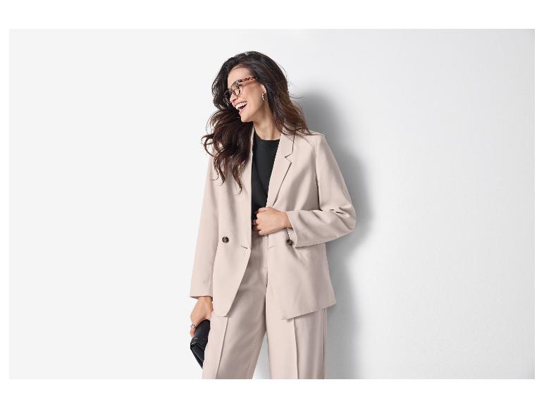 Frau in hellem Blazer und Hose, schwarzem T-Shirt und Brille, lächelnd zur Seite.