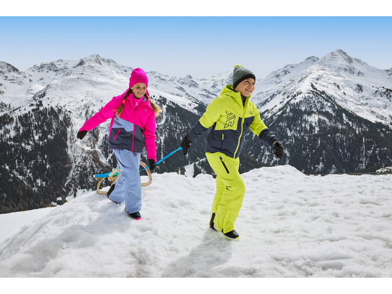 Zwei Kinder in bunten Skianzügen und Mützen, die einen Schlitten in einer verschneiten Berglandschaft ziehen.