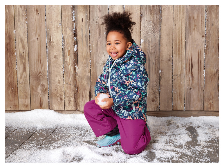 Ein Mädchen mit einer Winterjacke mit Blumenmuster und rosa Hose spielt im Schnee.