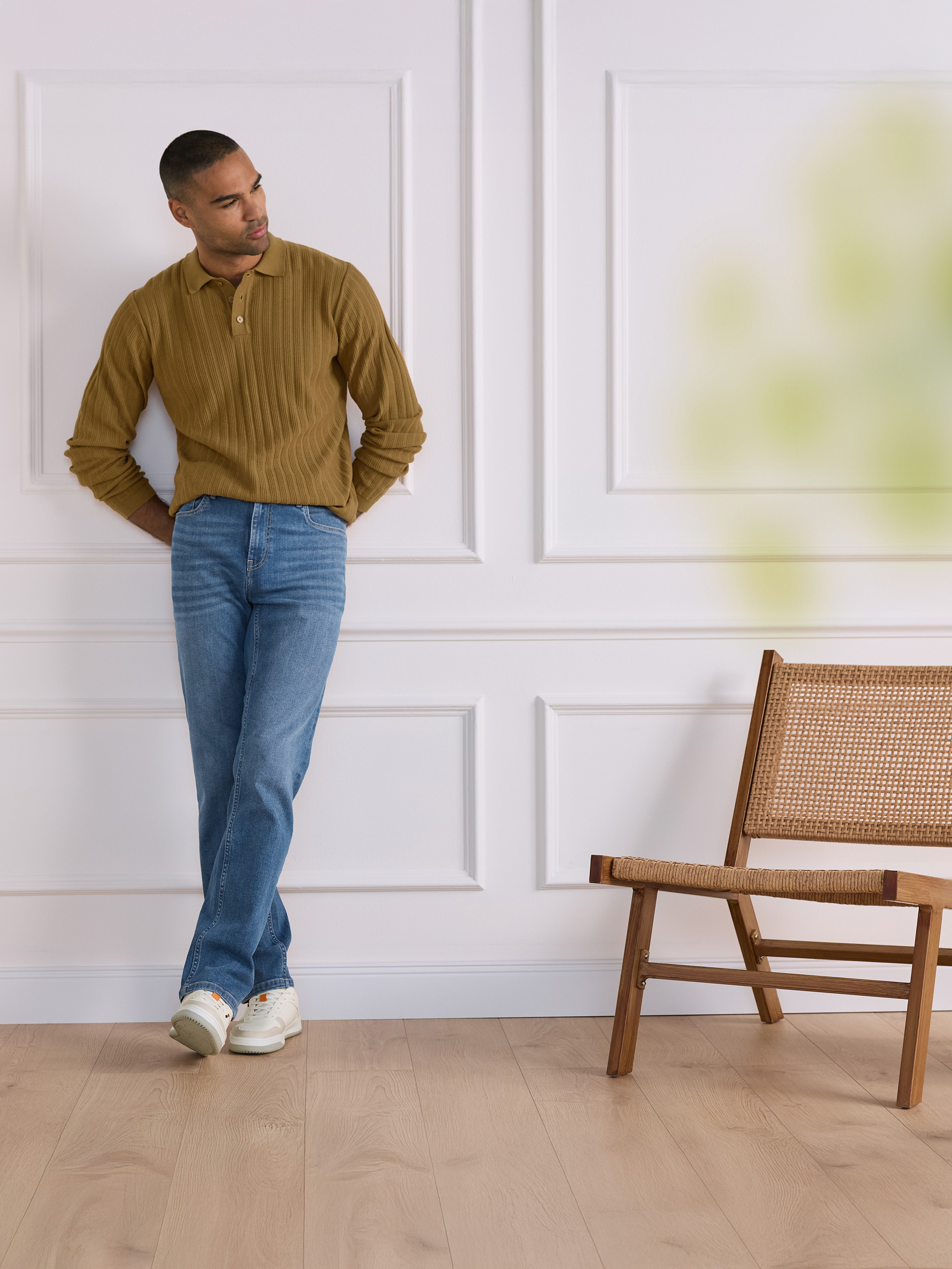 Mann in braunem geripptem Poloshirt und Jeans lehnt an einer weißen Wand mit Holzstuhl.