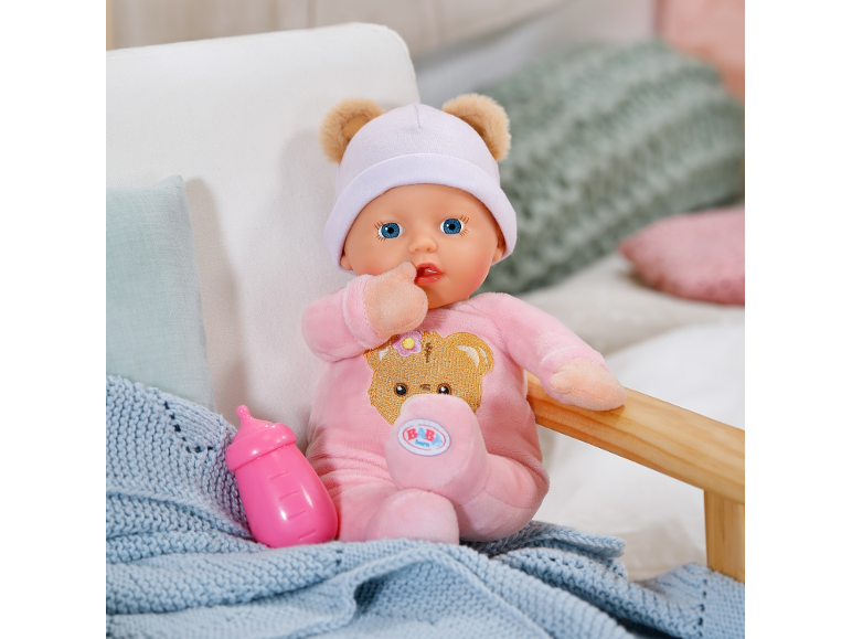 Weiche Babypuppe im rosa Strampler und Mütze mit Bärenohren neben einem Fläschchen.