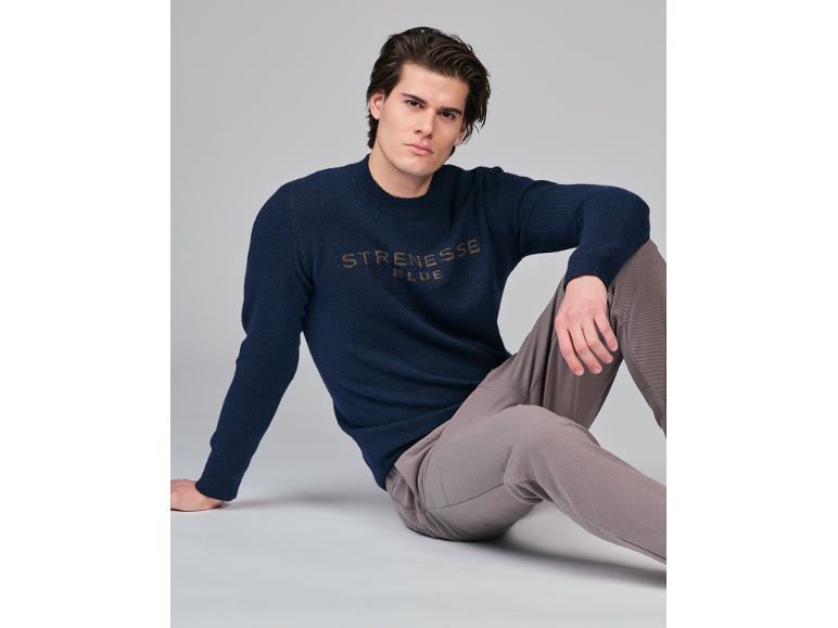 Mann in dunkelblauem Pullover mit Schriftzug und grauer Hose, sitzend auf grauem Hintergrund.