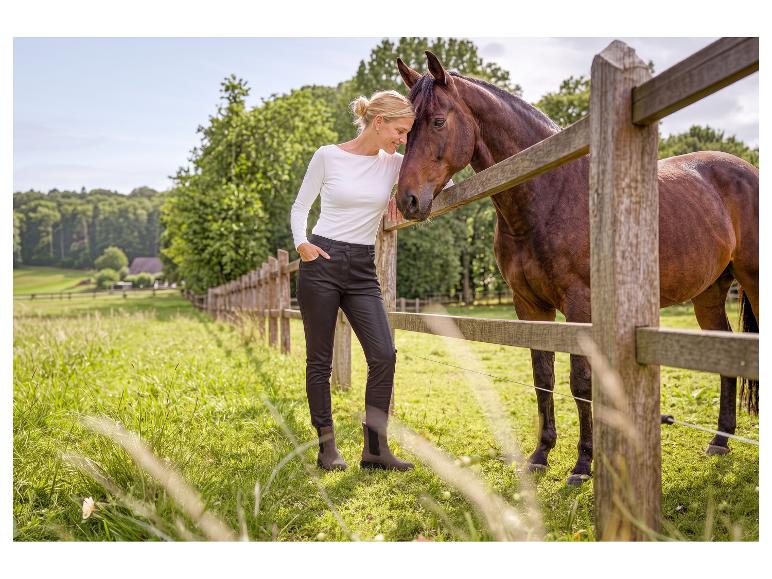 Frau in weißem Shirt und dunkler Hose mit Pferd an Holzzaun in ländlicher Landschaft.