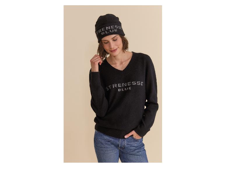Eine Frau in einem schwarzen Pullover und einer Mütze mit dem Text „STRENESSE BLUE“, die blaue Jeans trägt.