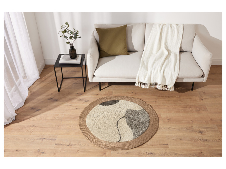 Ein runder Jute-Teppich mit abstraktem Muster vor einem weißen Sofa mit einer weißen Decke.