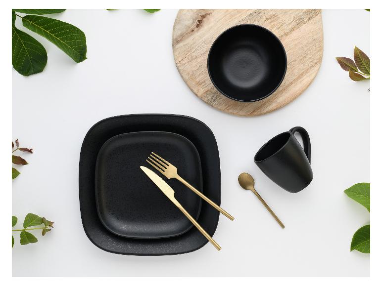 Schwarzes Geschirr mit goldenem Besteck, Schale und Tasse auf Holz und weißem Hintergrund mit Blättern.