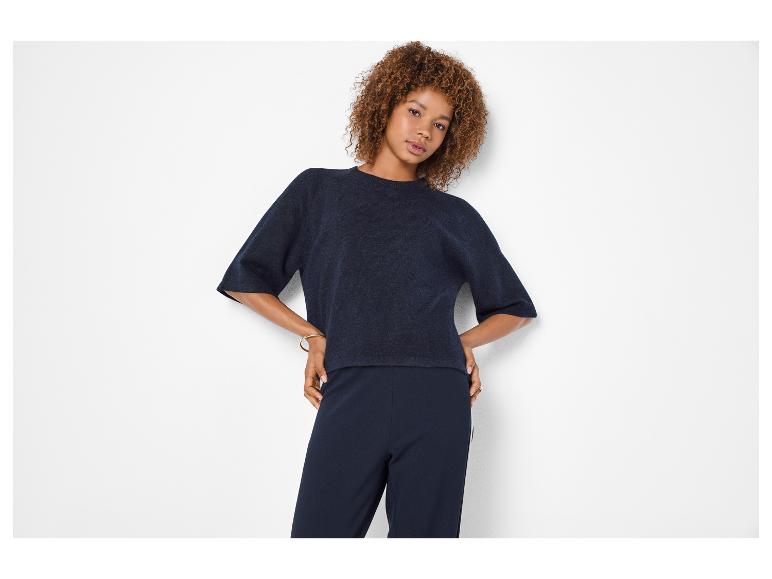 Frau mit lockigem Haar, trägt einen dunkelblauen Kurzarmpullover und dunkelblaue Hose.