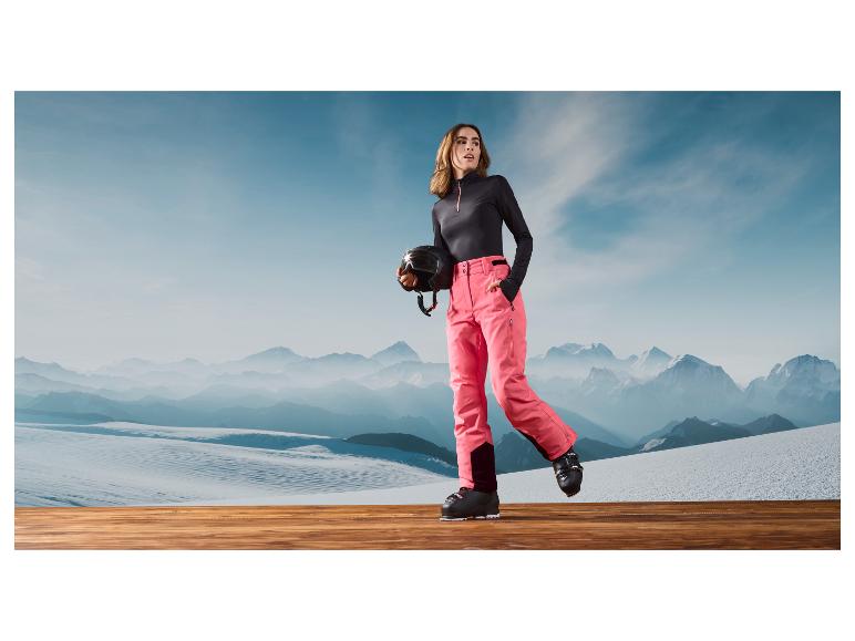 Eine Frau in rosa Skihose, schwarzen Skischuhen und Helm vor einer Berglandschaft.
