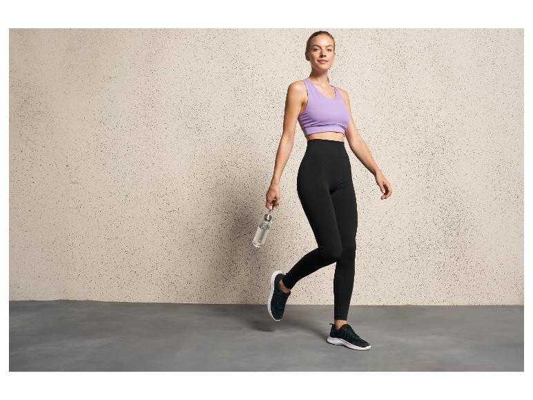 Eine Frau in schwarzen Leggings und einem lila Sport-BH posiert mit einer Wasserflasche.