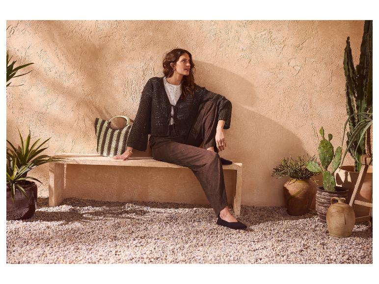 Frau in brauner Strickjacke und weiter brauner Hose, auf einer Steinbank mit Pflanzen sitzend.
