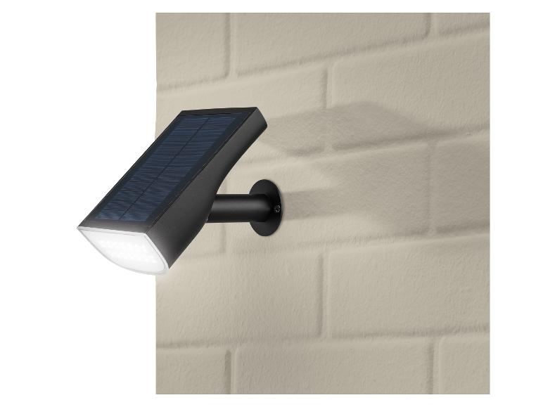 Schwarze LED-Solarleuchte mit Solarpanel, an einer beigen Ziegelwand montiert.