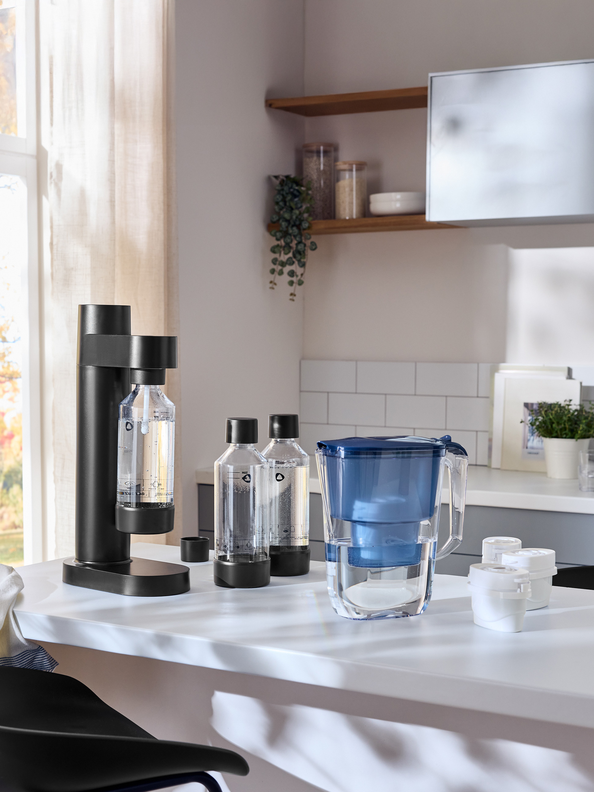 Schwarzer Sprudler mit Flaschen und einem blauen Wasserfilterkrug auf einer weißen Arbeitsplatte.