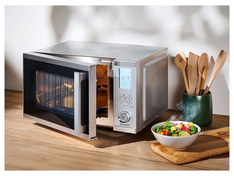 Silberne Mikrowelle mit offener Tür und einer Schüssel Gemüse auf einem Holzbrett.