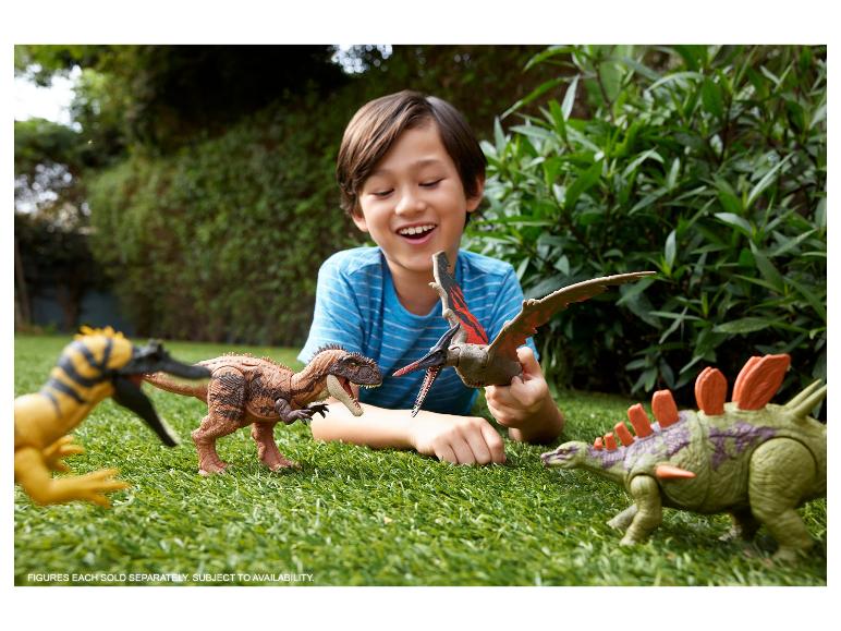 Ein Junge spielt mit Dinosaurier-Spielzeug auf dem Rasen, mit dem Text: „FIGUREN WERDEN EINZELN VERKAUFT. VERFÜGBARKEIT VORBEHALTEN.“