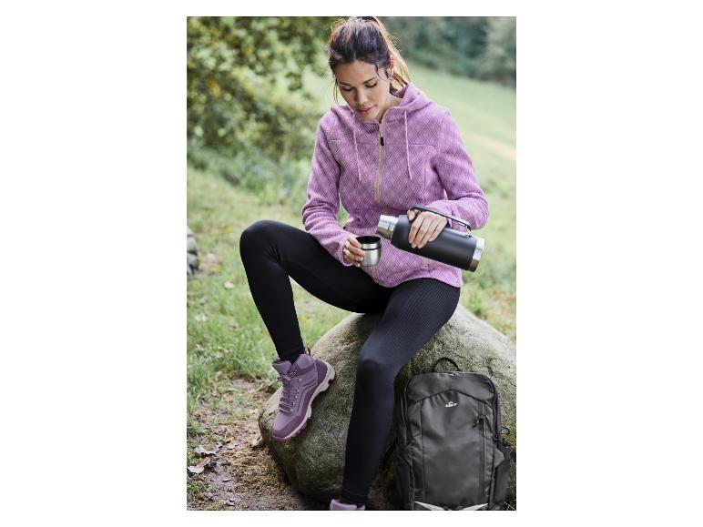 Eine Frau in einem rosa Hoodie, schwarzen Leggings und Wanderschuhen mit einer Thermoskanne und einem Crivit-Rucksack.