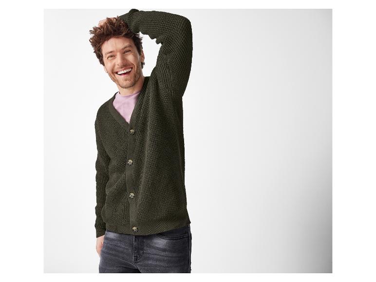 Ein lächelnder Mann in einer dunkelgrünen Strickjacke und dunklen Jeans.