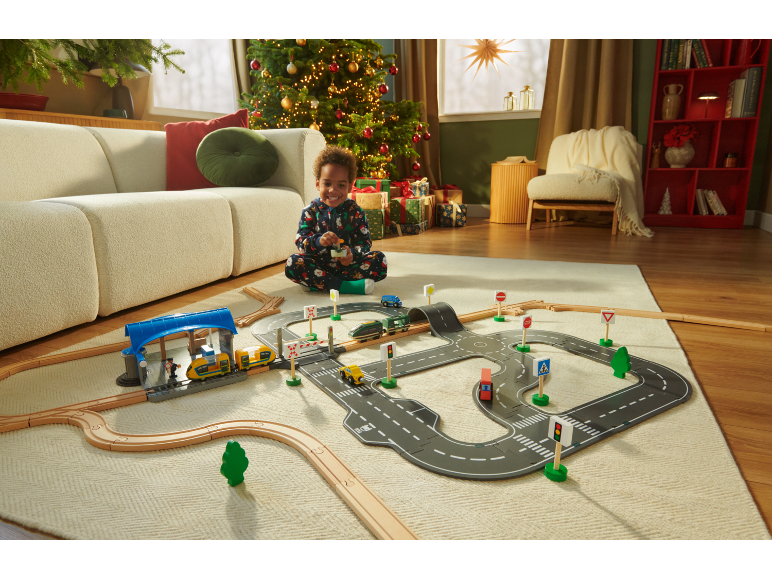 Kind spielt mit Holzeisenbahn und Straßenmatte auf einem Teppich in einem festlichen Wohnzimmer.