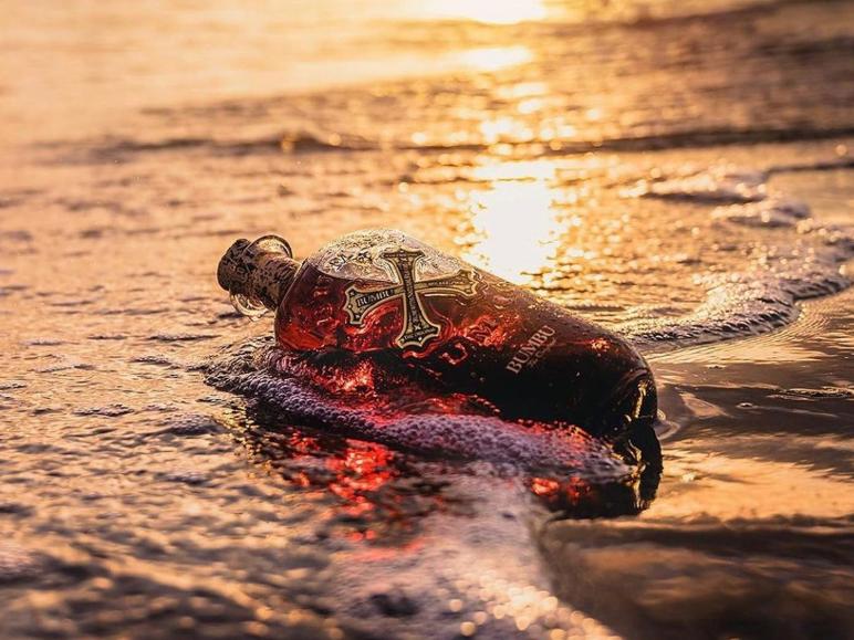 Flasche bernsteinfarbener Rum am Ufer bei Sonnenuntergang, mit schäumenden Wellen.