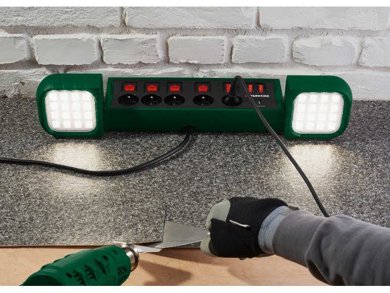 Steckdosenleiste mit integrierten LED-Leuchten, die eine Person beleuchten, die Bodenbelag mit einem Heißluftgebläse entfernt.