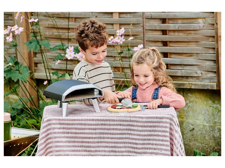 Zwei Kinder spielen mit einem Spielzeug-Pizzaofen und schneiden eine Spielzeugpizza draußen.