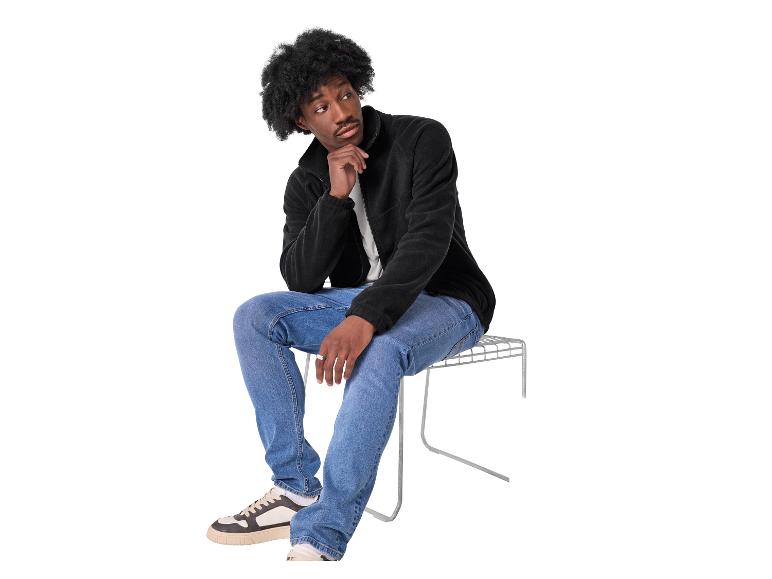 Ein Mann in schwarzer Fleecejacke und blauen Jeans sitzt nachdenklich auf einem Metallhocker.
