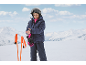 Kind in Skijacke, Skihose, Helm und Handschuhen, das einen Handschuh mit Skistöcken in der Nähe anpasst.