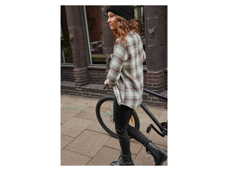 Eine Frau in einem karierten Hemd, schwarzen Hosen und Stiefeln geht an einem Fahrrad vorbei.