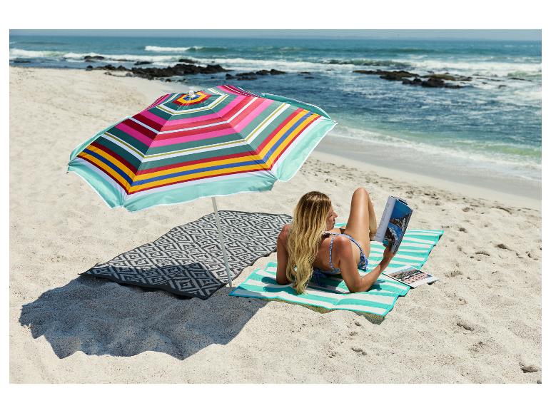 Frau liest auf einer CRIVIT Strandmatte unter einem bunten Sonnenschirm am Strand.