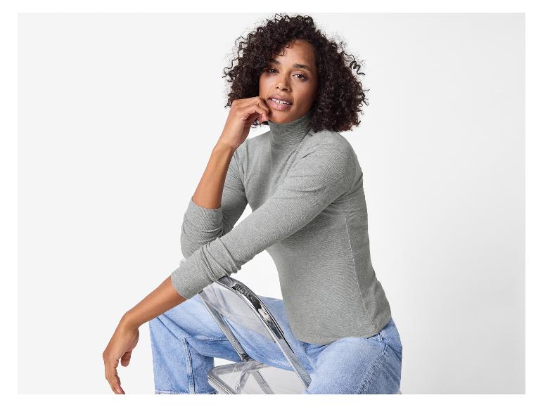 Frau in grauem Rollkragenpullover und Jeans sitzt auf einem transparenten Stuhl.