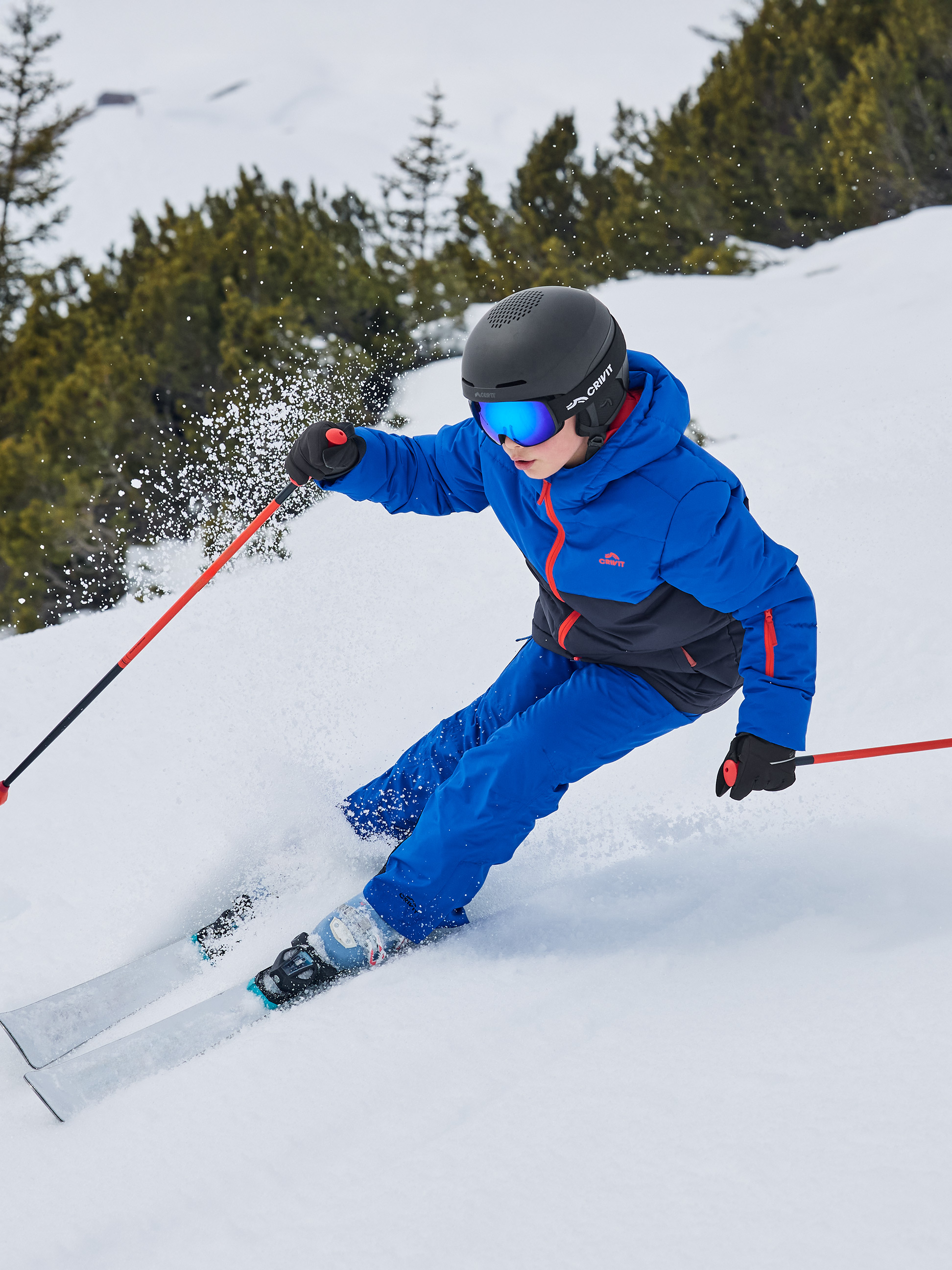 Kind in blauer Skijacke und -hose, Helm und Skibrille fährt einen verschneiten Hang hinunter.
