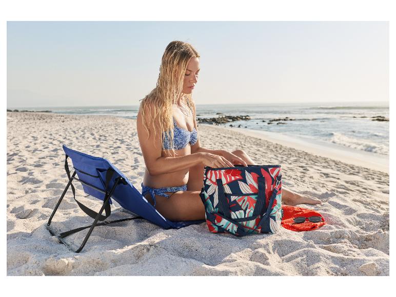 Frau im Bikini am Strand, sitzt neben einem blauen Strandstuhl und einer Tasche mit Tropenmuster.