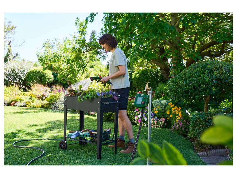 Frau gießt Pflanzen in einem Hochbeet mit einem Schlauch, neben einer Bewässerungsuhr.