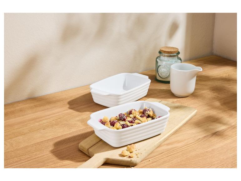 Zwei weiße Auflaufformen, eine mit Beeren-Crumble-Dessert, auf einem Holzbrett.