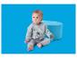 Baby in grauem Outfit mit Baufahrzeugen, sitzt neben einem blauen Pouf und hält eine Rassel.