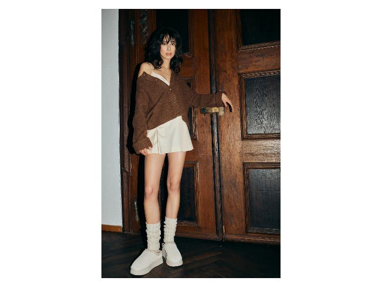 Frau in braunem Cardigan, weißem Top und plissiertem Minirock, mit weißen Hausschuhen und Socken.