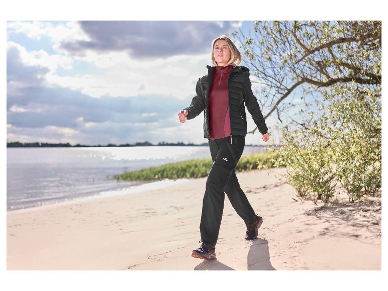 Frau in Outdoor-Jacke und -Hose spaziert am Sandstrand.