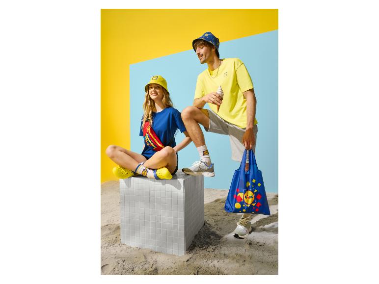 Zwei Personen in Sommerkleidung, Hüten, mit einer Tasche und Eiscreme vor Sandhintergrund.