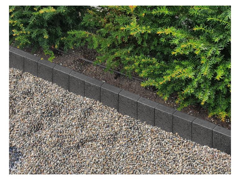 Graue Gartenbegrenzung trennt ein Kiesbeet und grüne Sträucher.