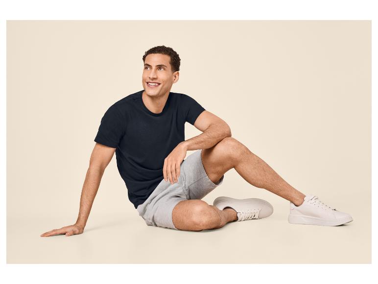 Mann in schwarzem T-Shirt und grauen Shorts sitzt auf dem Boden, lächelt und schaut nach oben.