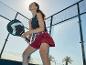 Frau in schwarzem Sporttop und rotem Rock mit Padel-Schläger auf dem Padel-Platz.