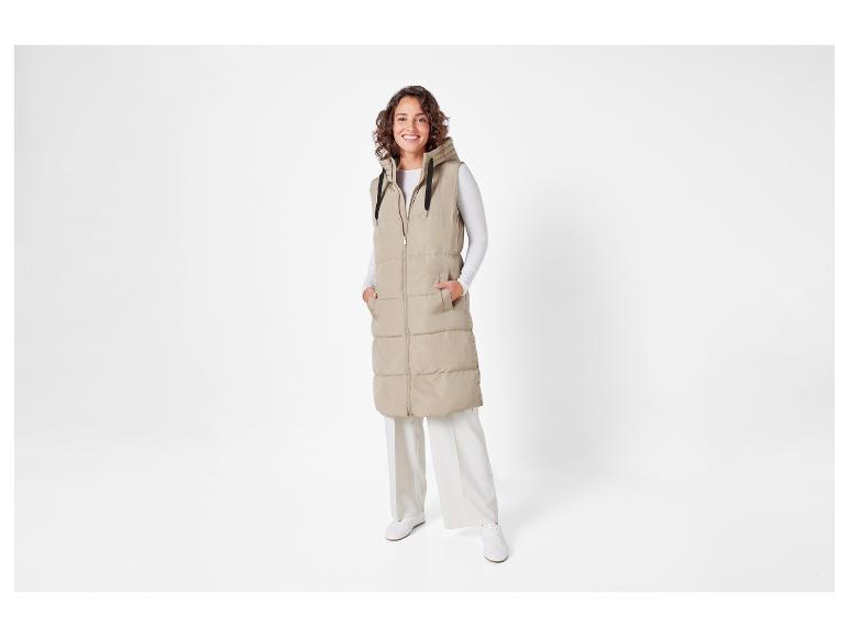 Eine Frau in einer beige wattierten Weste mit Kapuze und weißer Hose.