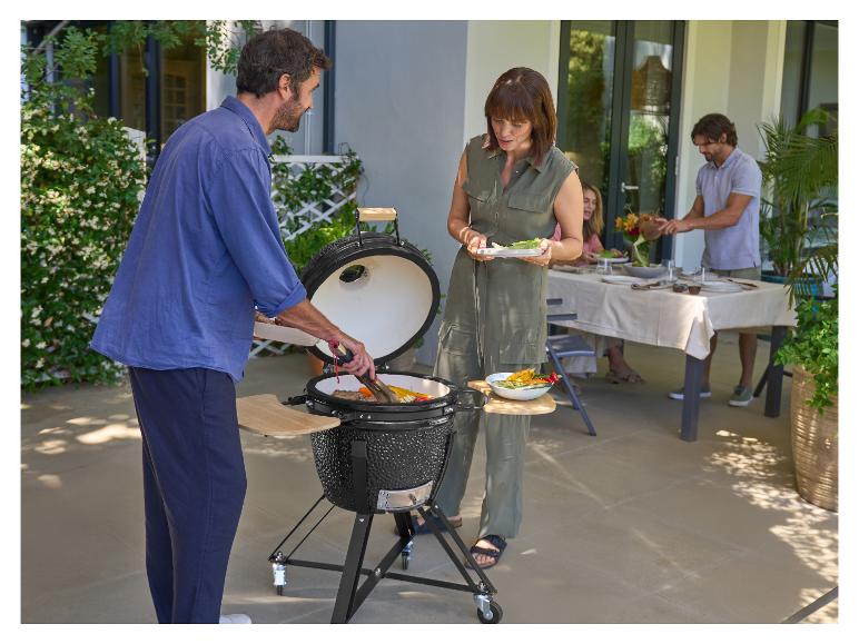 Leute grillen Essen auf einem Keramikgrill auf einer Terrasse, mit einem Esstisch im Hintergrund.