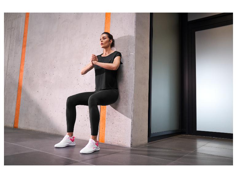 Frau in schwarzer Sportkleidung und weißen Sportschuhen macht einen Wall Sit.