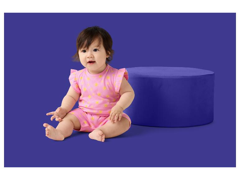 Ein Baby in einem rosa Strampler mit Sonnenmuster sitzt neben einem blauen Pouf.