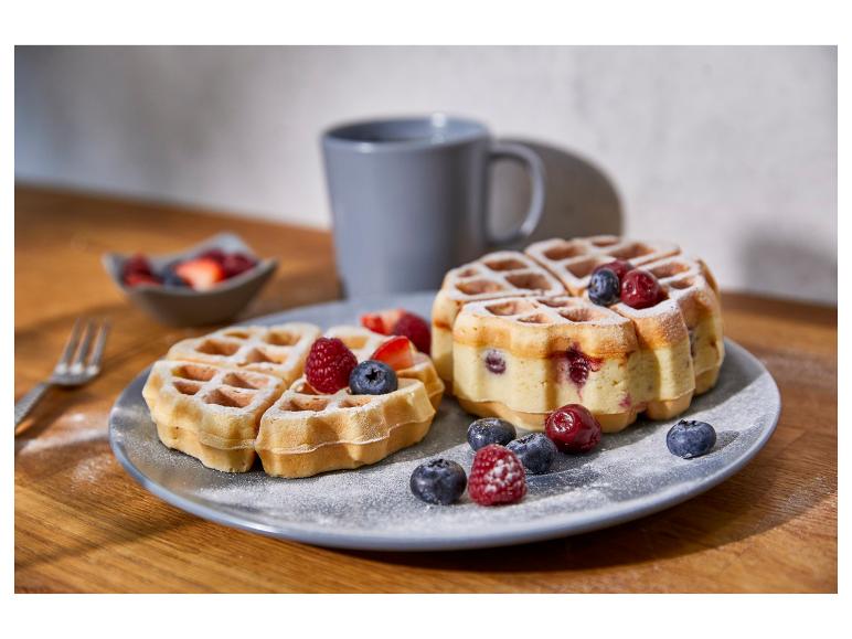 Waffeln mit Beeren und Puderzucker auf einem Teller, mit einer Kaffeetasse im Hintergrund.