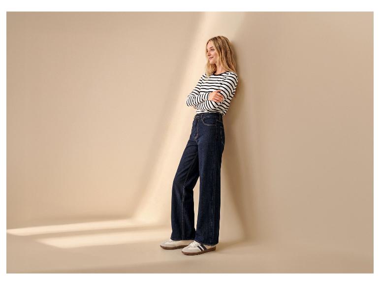 Frau in gestreiftem Shirt und weiter Jeans, an eine Wand gelehnt, trägt Sneaker.