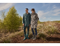 Mann und Frau in Regenmänteln und Stiefeln in der Natur, mit Bäumen und blauem Himmel im Hintergrund.