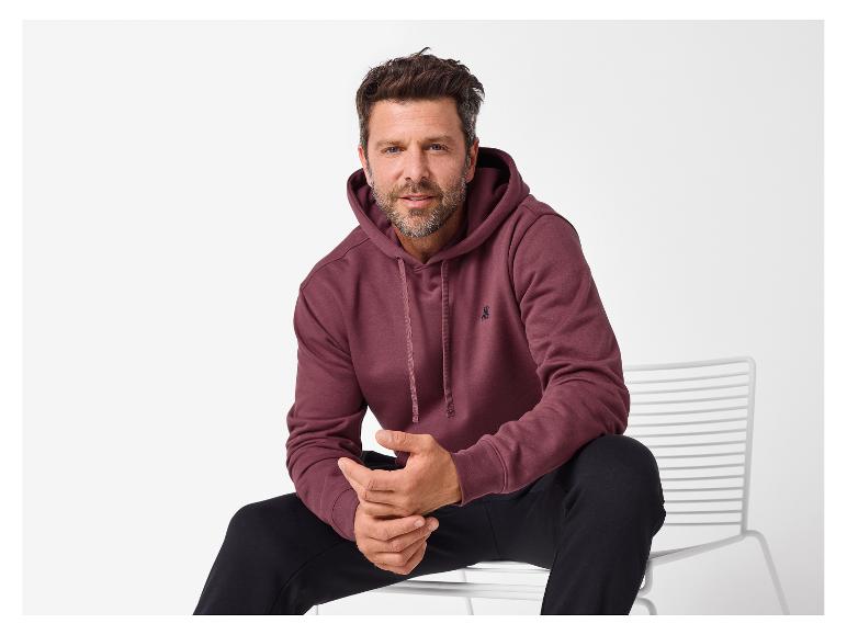 Mann in einem bordeauxroten Kapuzenpullover und schwarzer Hose sitzt auf einem weißen Stuhl.