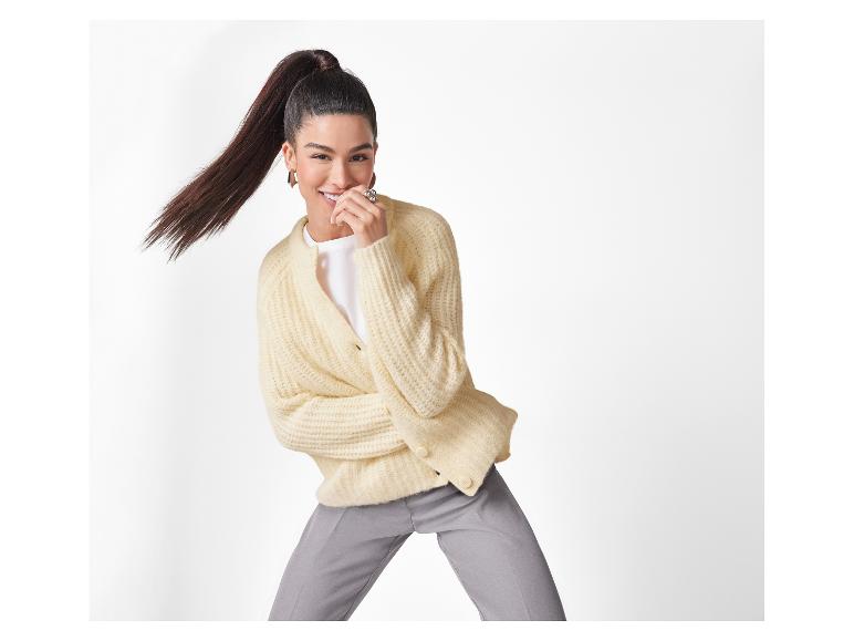 Lächelnde Frau mit Pferdeschwanz, trägt eine cremefarbene Strickjacke und graue Hose.