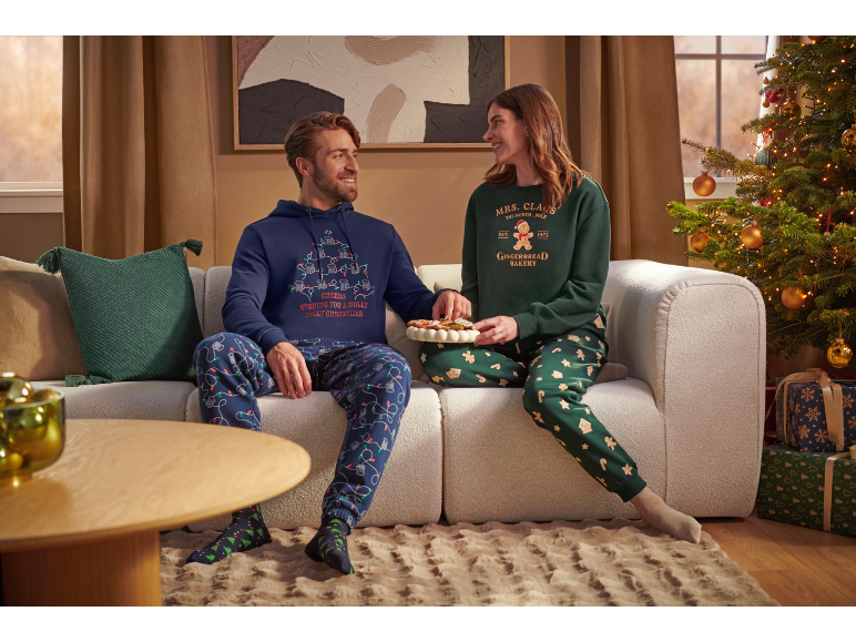 Mann und Frau in weihnachtlichen Pyjamas sitzen auf einem Sofa neben einem Weihnachtsbaum.